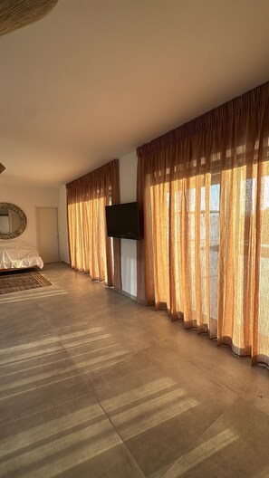 Una habitación amplia con una cama grande, un televisor de pantalla plana en la pared y un ventanal con cortinas traslúcidas.
