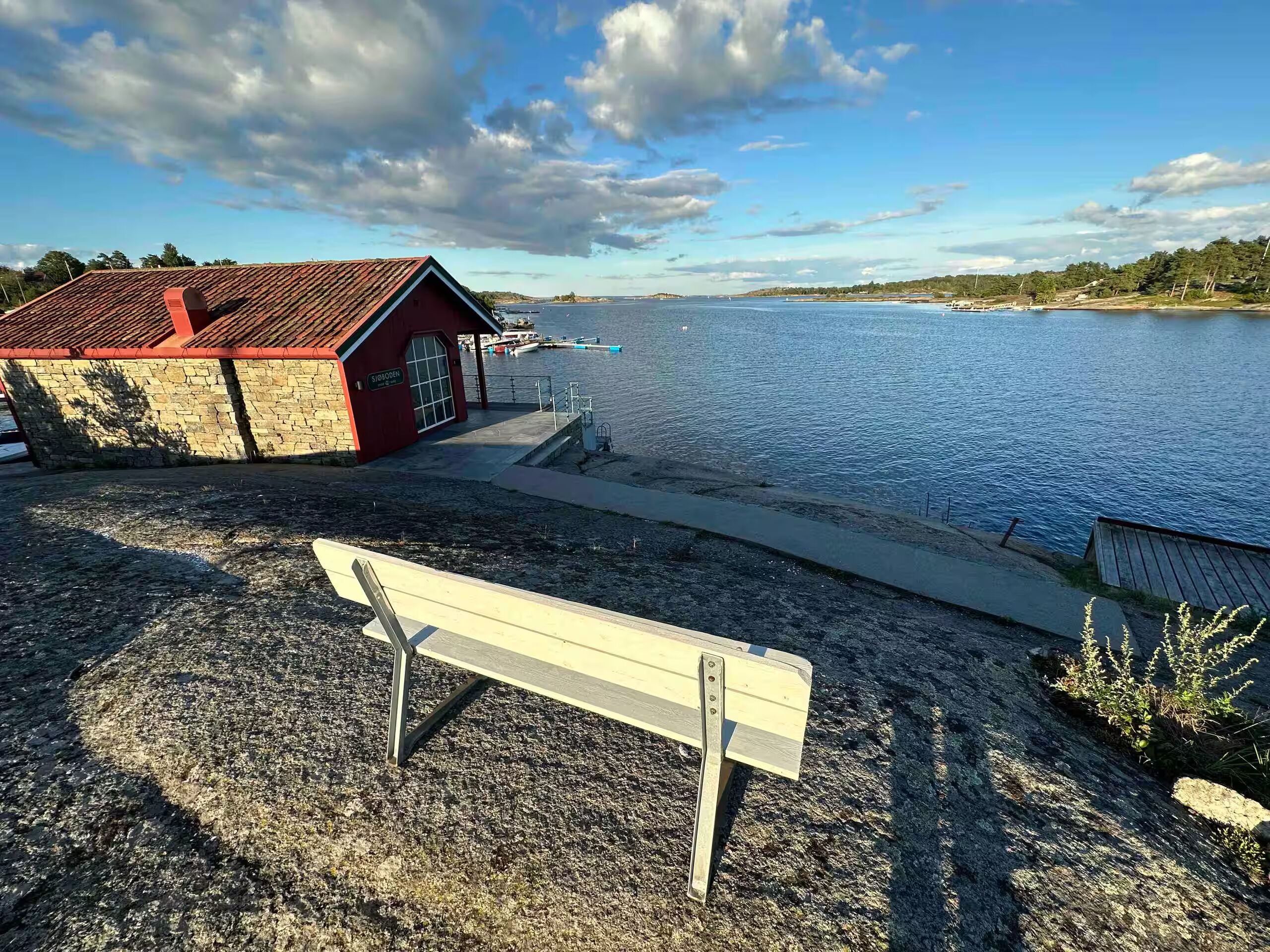 Une vue sur la côte, avec un bâtiment rouge portant l’inscription « SJOODEN » et un banc blanc sur une plage de galets.