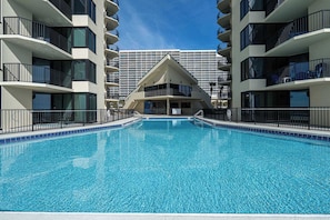 A swimming pool with a clear blue water surface, surrounded by a low fence, and a building with multiple balconies in the background. - Coastal Bliss and Beachfront Resort Oasis for 5! (Panama City Beach)