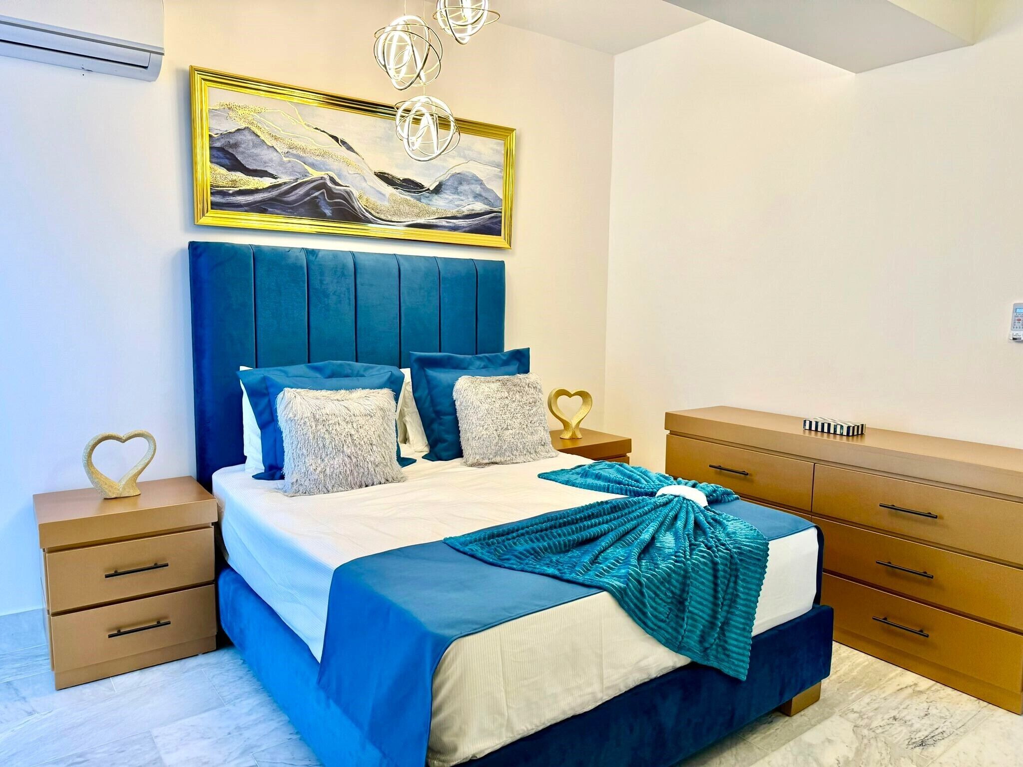 Un dormitorio con cama, mesitas de noche, cómoda, un cuadro y lámpara en el techo.