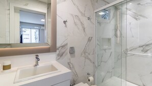 Un baño moderno con salpicadero de mármol, cabina de ducha de vidrio y tocador con lavamanos y espejo.