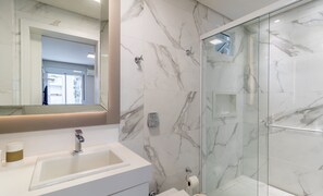 Ein modernes Badezimmer mit Marmorverkleidung, einer Glasdusche und einem Waschtisch mit Spüle und Spiegel.
