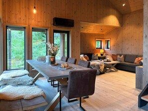 A spacious living area with a long dining table, comfortable seating, and large windows offering views of the outdoors.