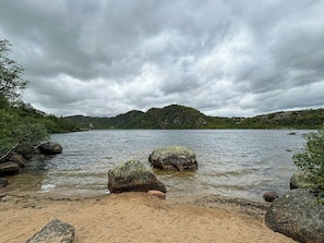 A river with rocks and a sandy beach, surrounded by hills and trees under a cloudy sky. - Mountain cabin in Ljosland with ski-in access (Åseral)