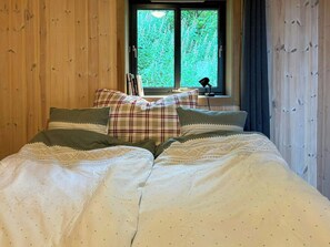A bed with a white duvet and patterned pillows, a window with a view of greenery, and a desk with a lamp. - Mountain cabin in Ljosland with ski-in access (Åseral)