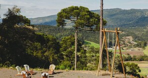 A rustic outdoor seating area with wooden chairs and a swing set, overlooking a lush valley with trees and distant hills.