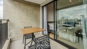 Ein Balkon mit Tisch und Stühlen, ein Bett mit weißer Bettdecke und Blick auf die Stadt durch ein großes Fenster.