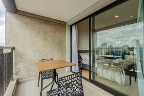 A balcony with a table and chairs, a bed with a white cover, and a city view through a large window. - Studio in Vila Cordeiro Ctb2505 (São Paulo)