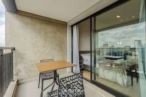 A balcony with a table and chairs, a bed with a white cover, and a city view through a large window. - Studio in Vila Cordeiro Ctb2505 (São Paulo)