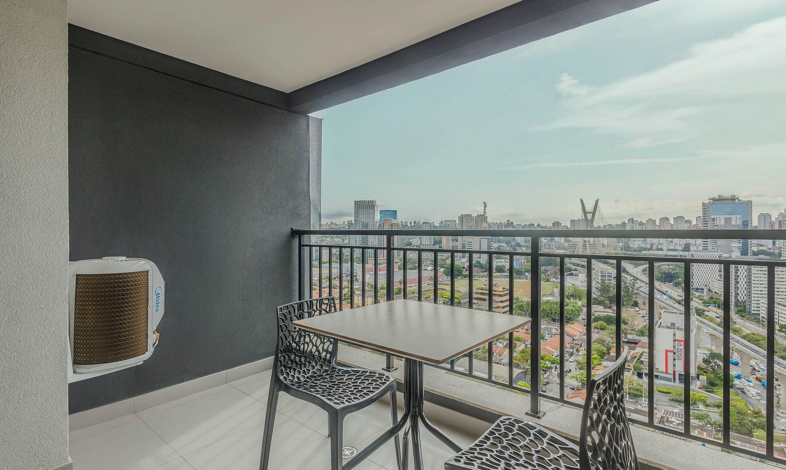 A balcony with a table and chairs, a view of a cityscape, and a building with a bridge in the distance.