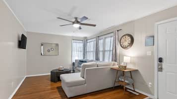 A room with a ceiling fan, a sofa, a side table with a lamp, a clock, and a television mounted on the wall.
