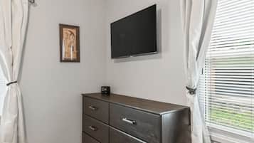 Et soveværelse med en mørk trækommode, et fladskærms-tv ophængt på væggen og et vindue med persienner.