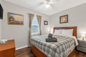 Une chambre à coucher comprenant un lit, une table de chevet, un bureau, un ventilateur de plafond et deux tableaux encadrés accrochés aux murs.