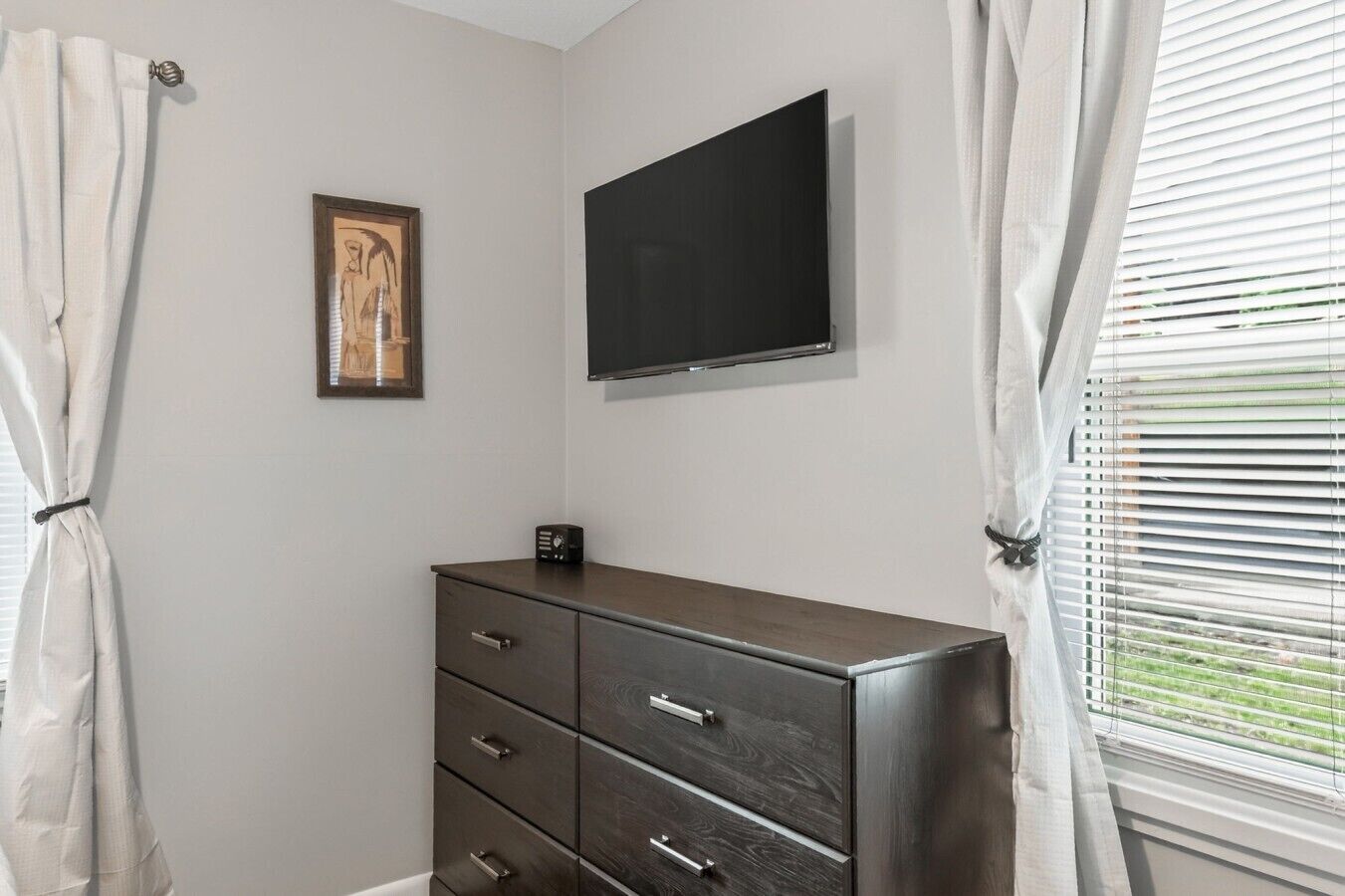 Un dormitorio con un aparador de madera oscura, un televisor de pantalla plana montado en la pared y una ventana con persianas.