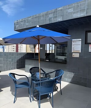 Un patio al aire libre con una mesa, sillas, un paraguas azul y un edificio al fondo.