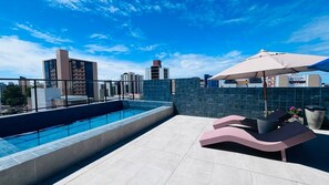 Un área de piscina en la azotea con vista despejada del horizonte de la ciudad, un espacio de relajación con tumbonas y un gran parasol que ofrece sombra.