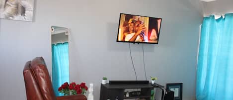 A living room with a television mounted on the wall, a leather armchair, a small table with decorative items, and a sofa.