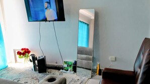 Una sala de estar con un televisor de pantalla plana montado en la pared, un espejo grande, un sillón de cuero marrón, un sofá, una mesita con un jarrón de rosas rojas y una consola de videojuegos.