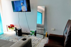 Una sala de estar con un televisor de pantalla plana montado en la pared, un espejo grande, un sillón de cuero marrón, un sofá, una mesita con un jarrón de rosas rojas y una consola de videojuegos.