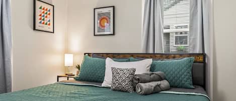 Una cama bien hecha con un edredón verde, una almohada estampada y una toalla enrollada. En la pared, dos obras de arte enmarcadas, una ventana con cortinas y una mesita de noche con una lámpara.