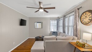 A modern living room with a ceiling fan, a sofa, a small table with a lamp, and a large wall clock. - 2Bdrm - 7 Minutes To Downtown (Kansas City)