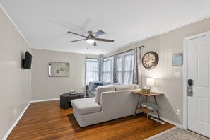 A room with a ceiling fan, a sofa, a side table with a lamp, a clock, and a television mounted on the wall.