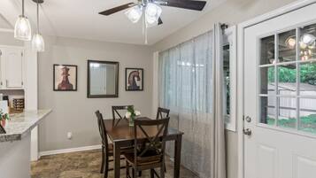 Uma área de refeições com mesa, cadeiras, ventilador de teto e janela com cortinas.
