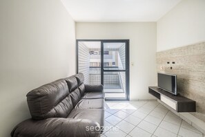 Una sala de estar moderna con un sofá de cuero, un televisor de pantalla plana sobre un mueble de madera y vistas a un balcón exterior a través de un ventanal.