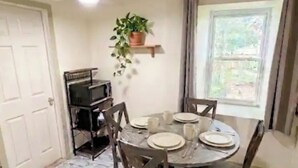 A dining area with a round table set for four, surrounded by wooden chairs. A ceiling fan is visible above, and a potted plant hangs on a wall-mounted shelf.