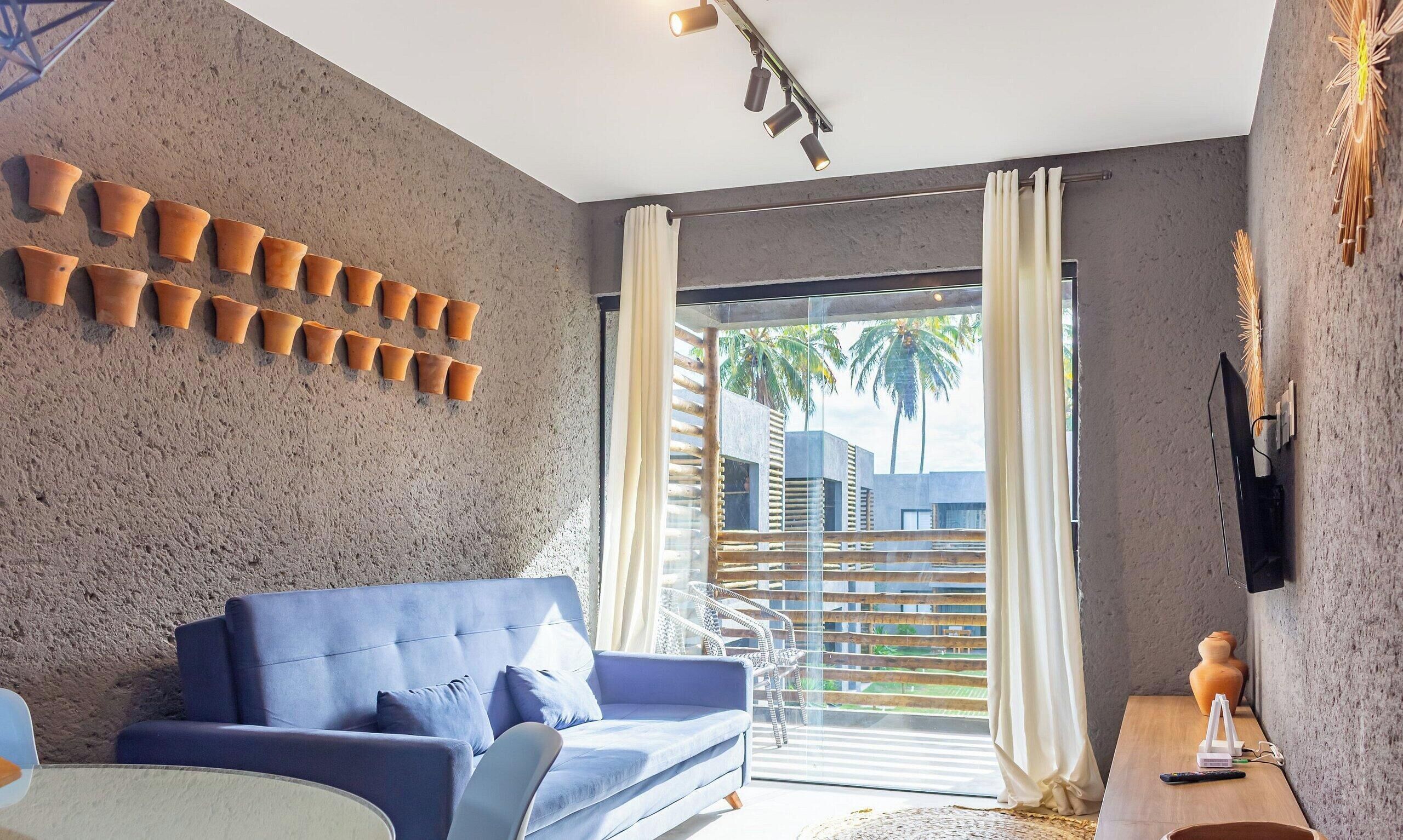 A modern living room with a blue sofa, a flat-screen TV mounted on the wall, and a large window with curtains.