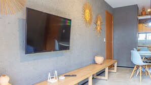 A modern living room with a flat-screen TV mounted on the wall, a wooden bench, a small table with a vase and remote control, and a dining area with a chair and table. - Nice Apart Near Patacho Beach Cvpa103 (Porto de Pedras)