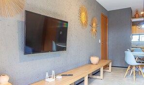 Ein modernes Wohnzimmer mit einem an der Wand befestigten Flachbildfernseher, einer Holzbank, einem kleinen Tisch mit Vase und Fernbedienung sowie einem Essbereich mit Stuhl und Tisch.