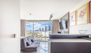 Uma sala de estar moderna com um sofá cinzento, uma televisão de tela plana fixada na parede e vista para a cidade através de grandes janelas.