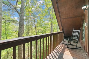 A wooden deck with a metal railing, a rocking chair, and a view of a forested area. - Marvelous 2-bedroom cabin located behind Dollywood. (Sevierville)