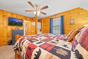 Ein Schlafzimmer im Holzstil, mit Deckenventilator, Fernseher, einem Bett mit geblümter Tagesdecke und einem Fenster mit Jalousien.
