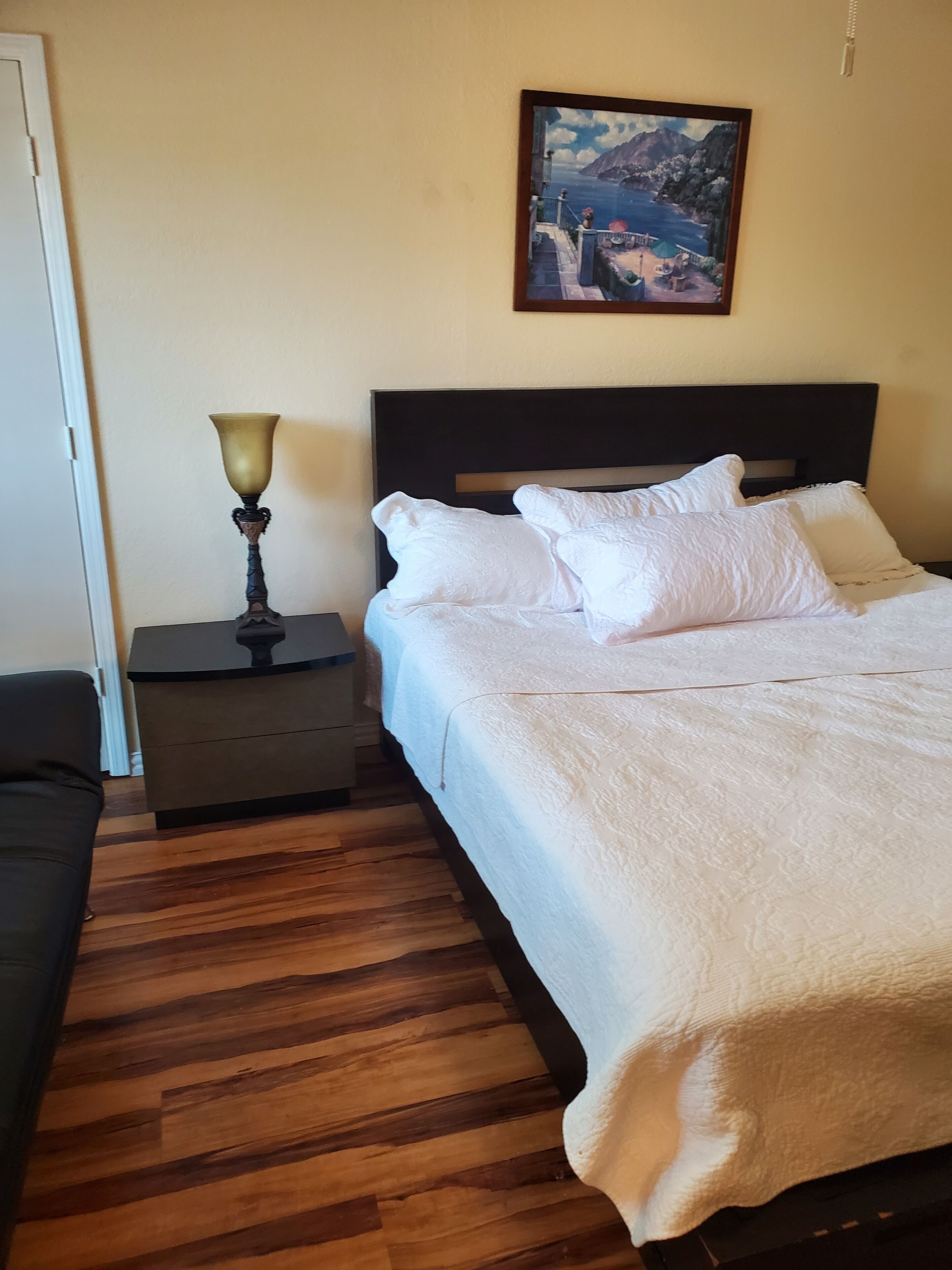 A neatly made bed with white bedding, a bedside table with a lamp, and a painting on the wall.