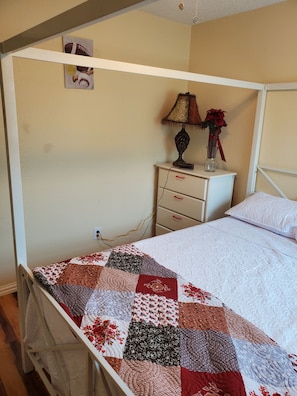Habitación individual con una cama y cabecera blanca, colcha con diseño de patchwork, mesita de noche con lámpara y un florero, además de un cuadro colgado en la pared.