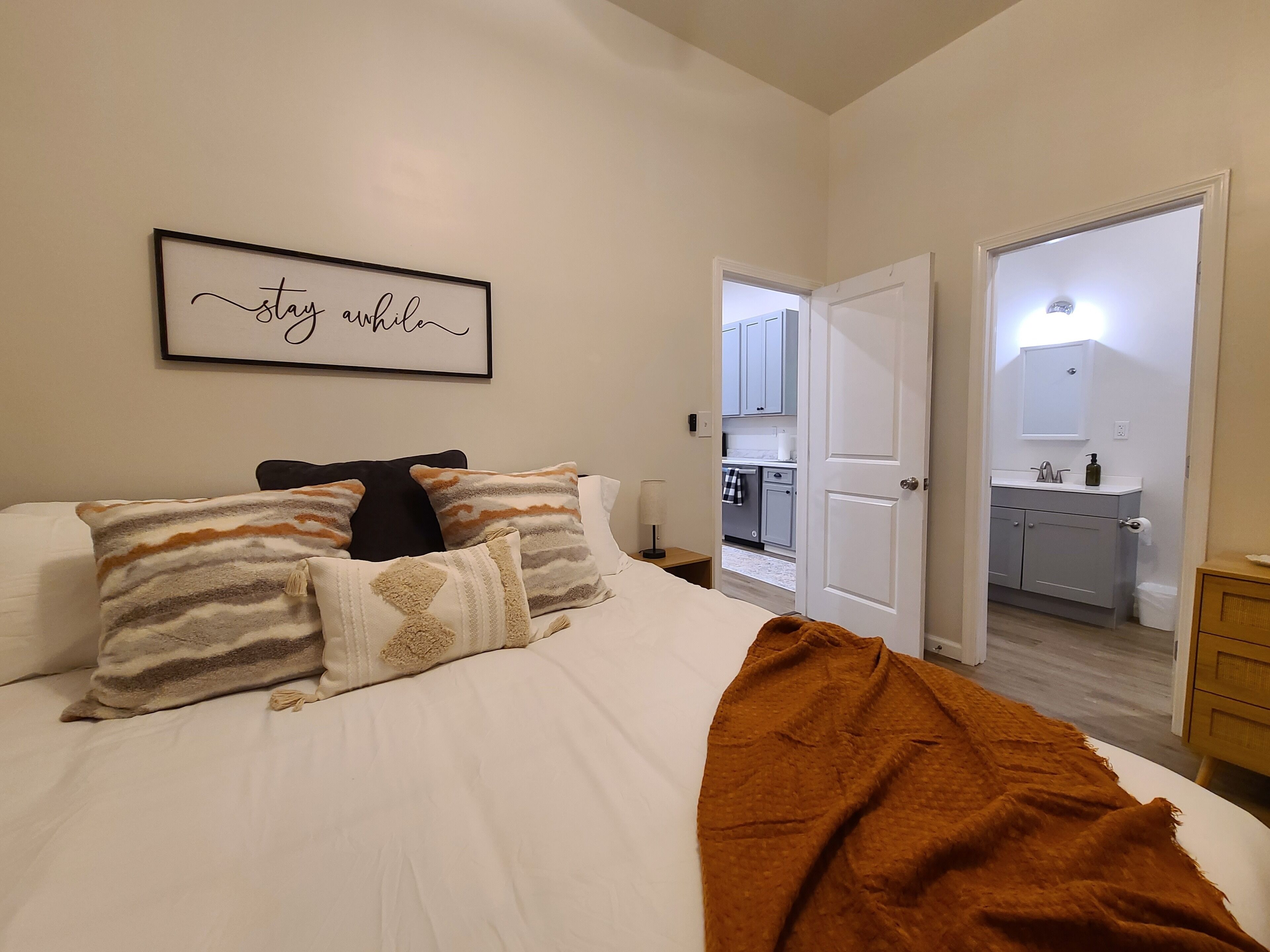 Un dormitorio con cama, almohadas, una colcha, una obra de arte en la pared y una puerta abierta que da a un baño.