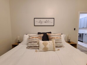 A neatly made bed with multiple pillows, a framed quote on the wall, and a visible kitchen area.