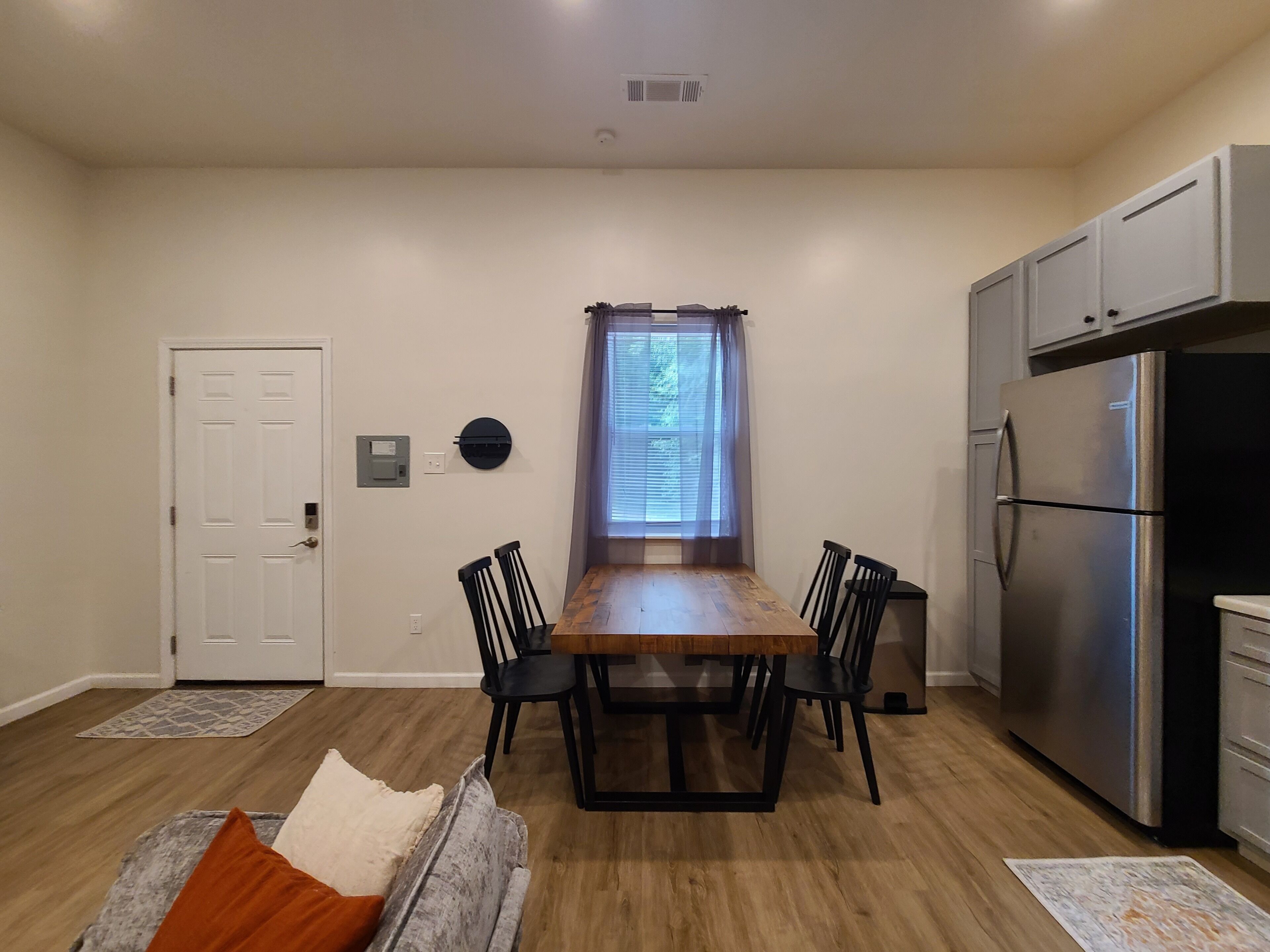 A dining area with a wooden table and chairs, a refrigerator, and a window with curtains.