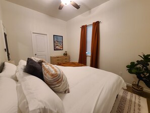 Une chambre à coucher avec un lit, une commode, une table de chevet, un tableau et une fenêtre avec des rideaux.