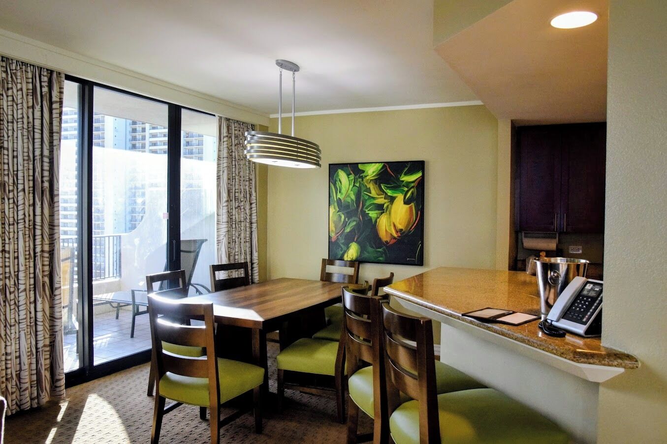 A hotel room with a dining area, a painting, a telephone, and a view of the city.