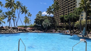 A hotel pool area with clear blue water, surrounded by palm trees and a large building with balconies. - Oceanfront View in Honolulu at The Hilton Hawaiian Village Lagoon Tower (Honolulu)