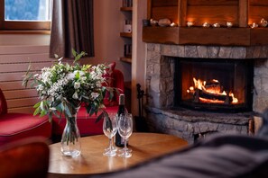 Ein gemütlicher Raum mit Kamin, einem Tisch mit Weingläsern und einer Blumenvase sowie einem roten Sessel.