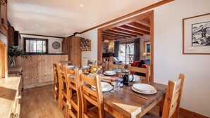 Un tavolo da pranzo in legno apparecchiato con piatti, bicchieri e una brocca, circondato da sedie in una stanza con pavimento in legno e soffitto spiovente.