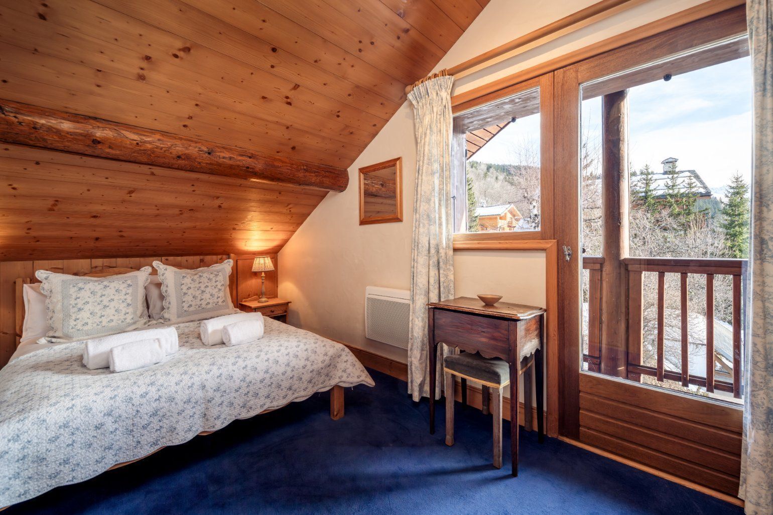 A bedroom with a wooden ceiling, a bed with floral bedding, a nightstand with a lamp, a small table, and a view through a glass door.