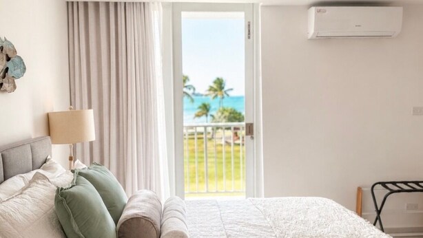 A hotel room with a bed, a window with curtains, a view of the ocean, and an air conditioning unit. - Tres Soles - Private Island Escape - Family Friendly - Isleta Marina (Fajardo)