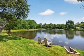 Ein friedlicher Blick auf einen See mit zwei Adirondack-Stühlen, die zum Wasser zeigen, umgeben von üppigem Grün und Bäumen.