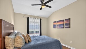 Quarto com cama, ventilador de teto, janela com cortinas e duas pinturas penduradas na parede.