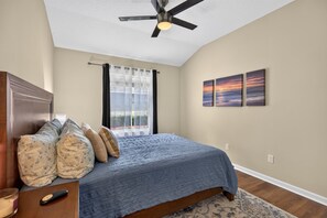 Ein Schlafzimmer mit einem Bett, einem Deckenventilator, einem Fenster mit Vorhängen und zwei an der Wand befestigten Bildern.